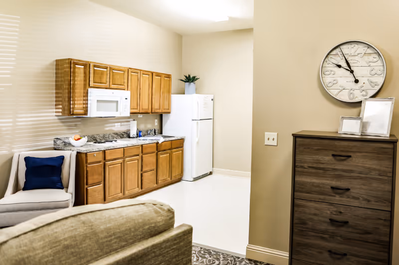 Interior view of a senior living facility kitchen area with wooden cabinets, a white microwave, a white refrigerator, a countertop with a sink, and a small potted plant. Adjacent to the kitchen is a beige wall with a round clock and a wooden chest of drawers with picture frames on top. A beige armchair with a dark blue cushion is partially visible in the foreground.