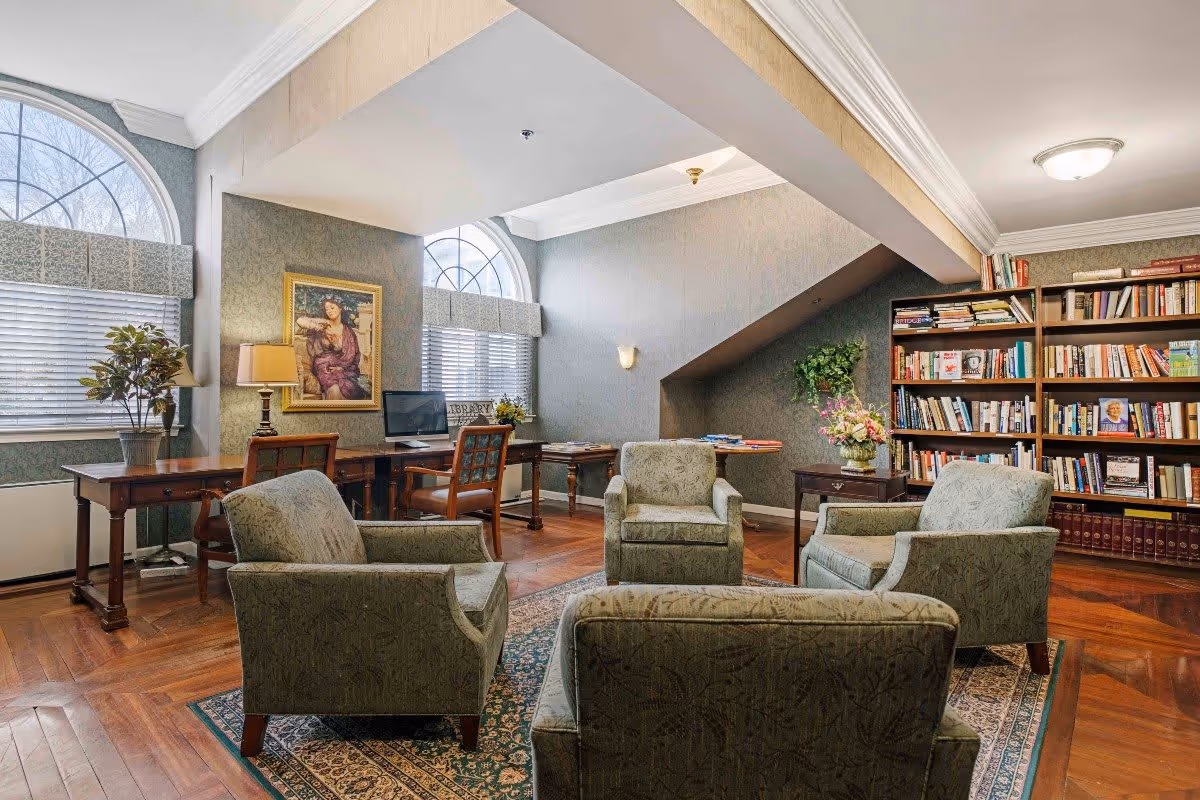 A cozy library or reading room with four upholstered armchairs arranged around a patterned rug. There are two wooden desks with chairs, a computer, a table lamp, and potted plants near two large arched windows with blinds. A bookshelf filled with books lines the right wall, and a framed painting hangs on the wall between the windows. The room has wooden flooring and soft lighting.