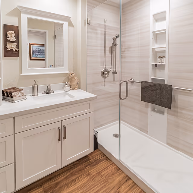 A clean and modern bathroom featuring a white vanity with a sink, a large mirror above it, and a glass-enclosed shower with a handheld showerhead and built-in shelves. The floor has wood-like flooring, and a dark towel hangs on the shower door handle. There are decorative items on the vanity and a small wall decoration beside the mirror.