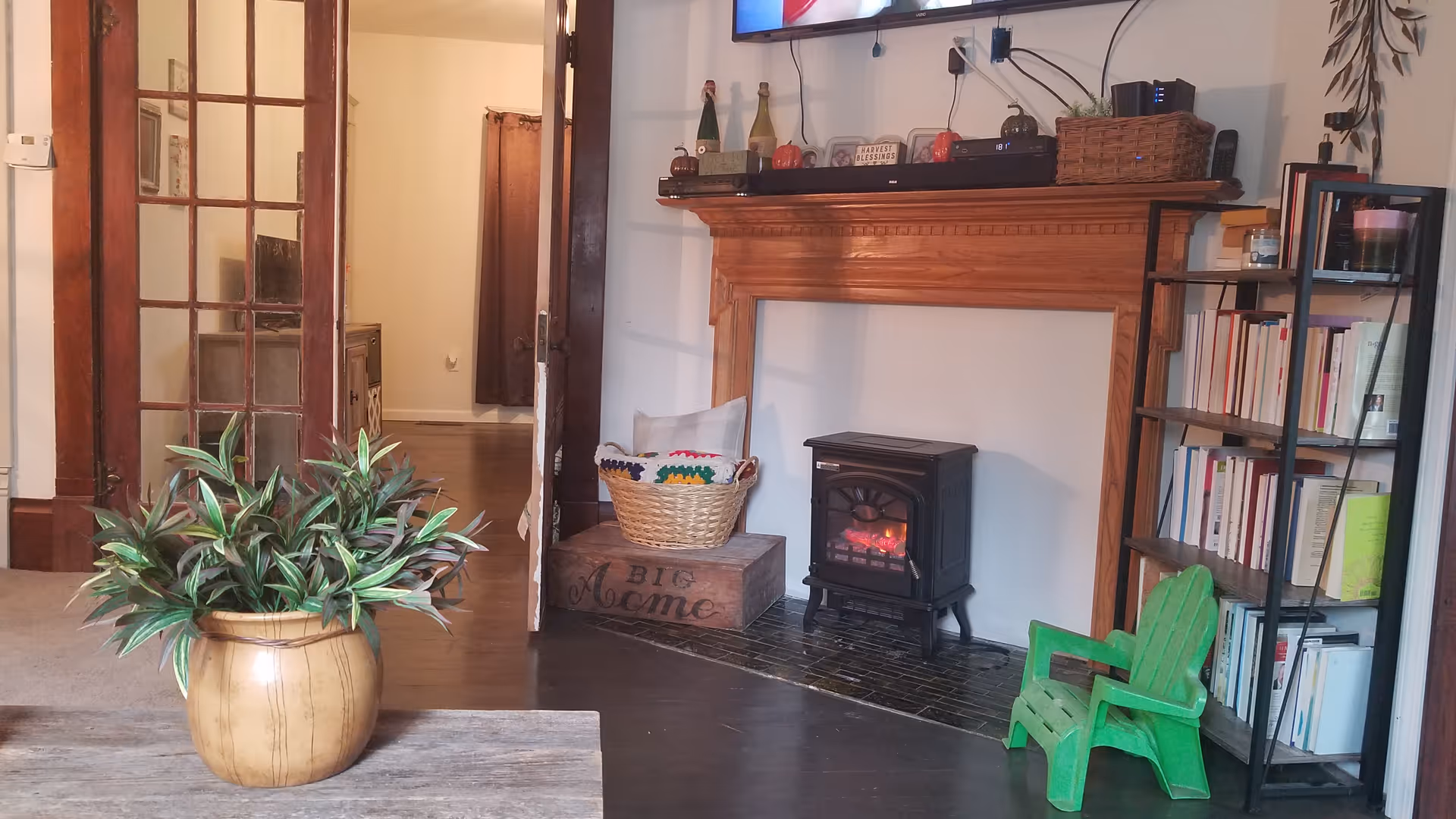 Cozy living room area with a wooden mantelpiece above a small electric fireplace. A green child-sized chair is placed next to a black metal bookshelf filled with books. A basket with pillows sits on a wooden crate labeled 'Big Acme.' A potted plant is on a wooden table in the foreground. French doors lead to another room with wooden flooring.