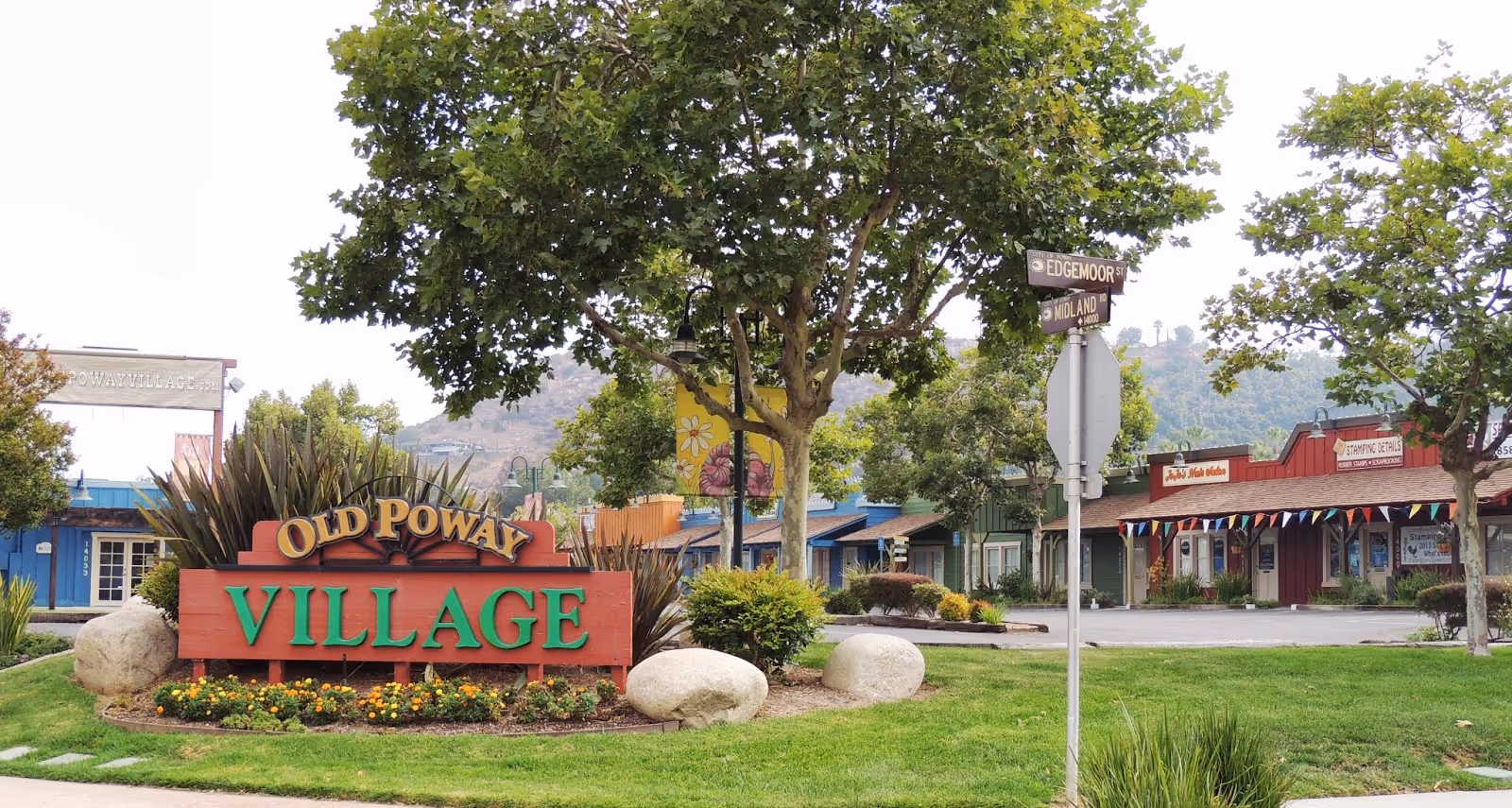 Outdoor view of a small village area with a large sign reading 'Old Poway Village' surrounded by greenery and flowers. There are trees, bushes, and a street sign indicating Edgemoor and Midland streets. Several small buildings with colorful facades and a mountain in the background are visible.