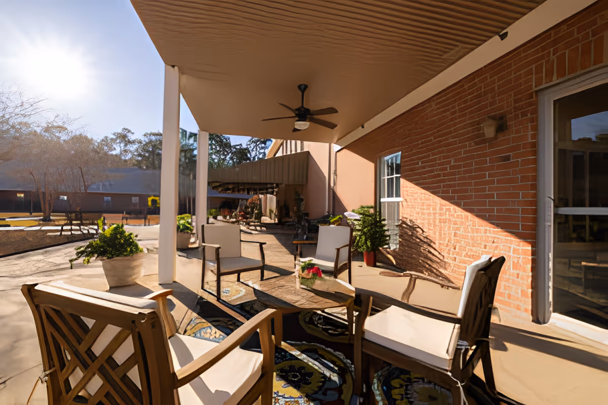 Covered outdoor patio area with cushioned chairs arranged around a glass-top coffee table, potted plants, a ceiling fan, and a brick wall with windows in the background. Sunlight casts shadows on the patio floor and wall.