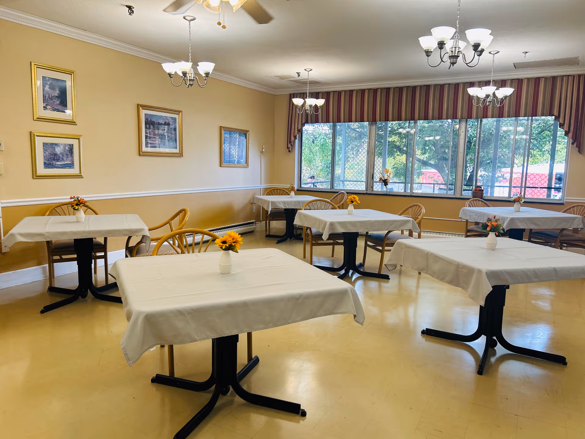 A dining room with several tables covered in white tablecloths, each adorned with a small vase of flowers. Wooden chairs are arranged around the tables. The room has beige walls decorated with framed pictures and large windows with striped valances letting in natural light. Ceiling fans and chandeliers provide lighting.