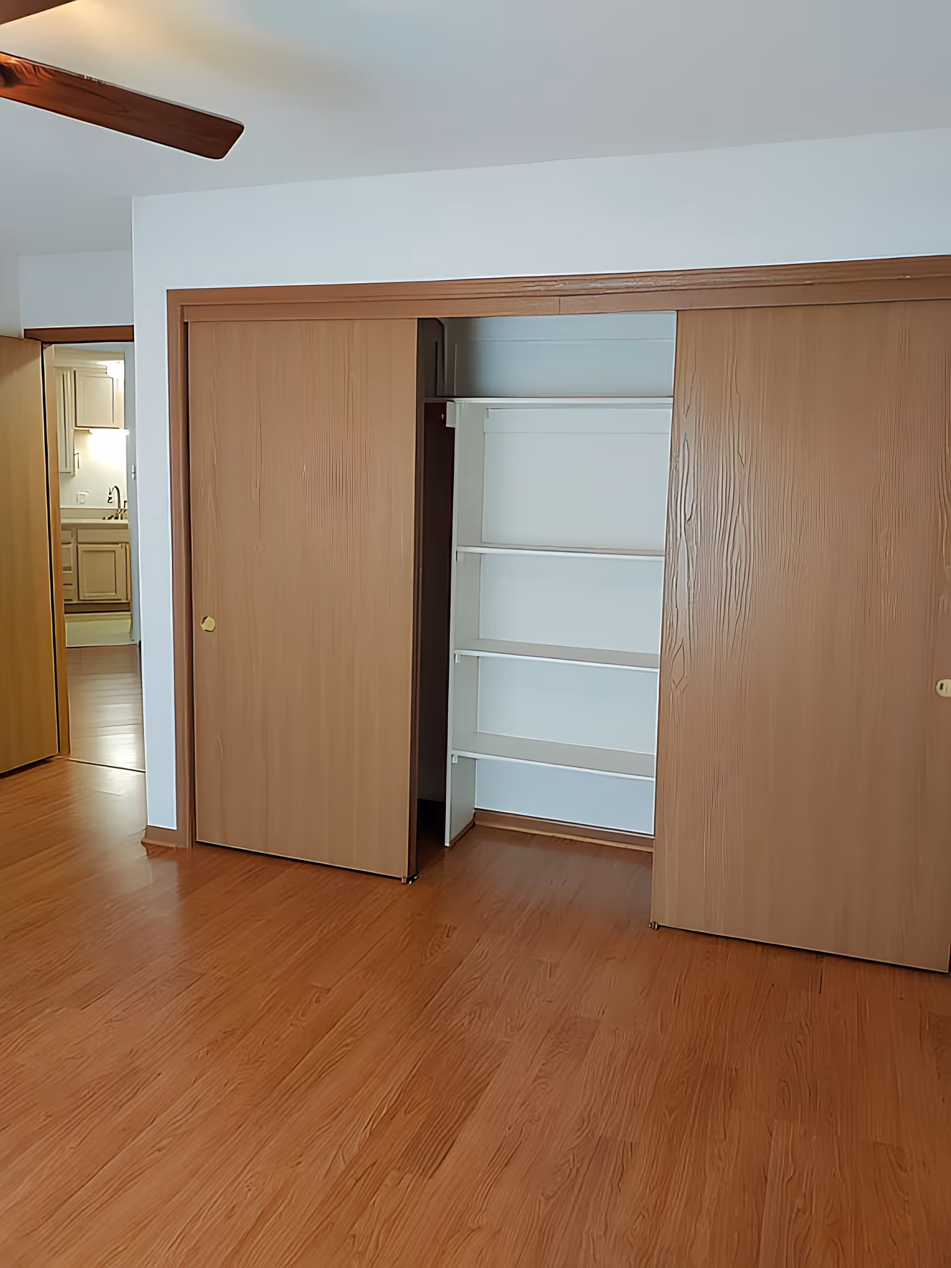 Empty room with hardwood floors, a ceiling fan, and sliding wooden closet doors revealing built-in shelves.