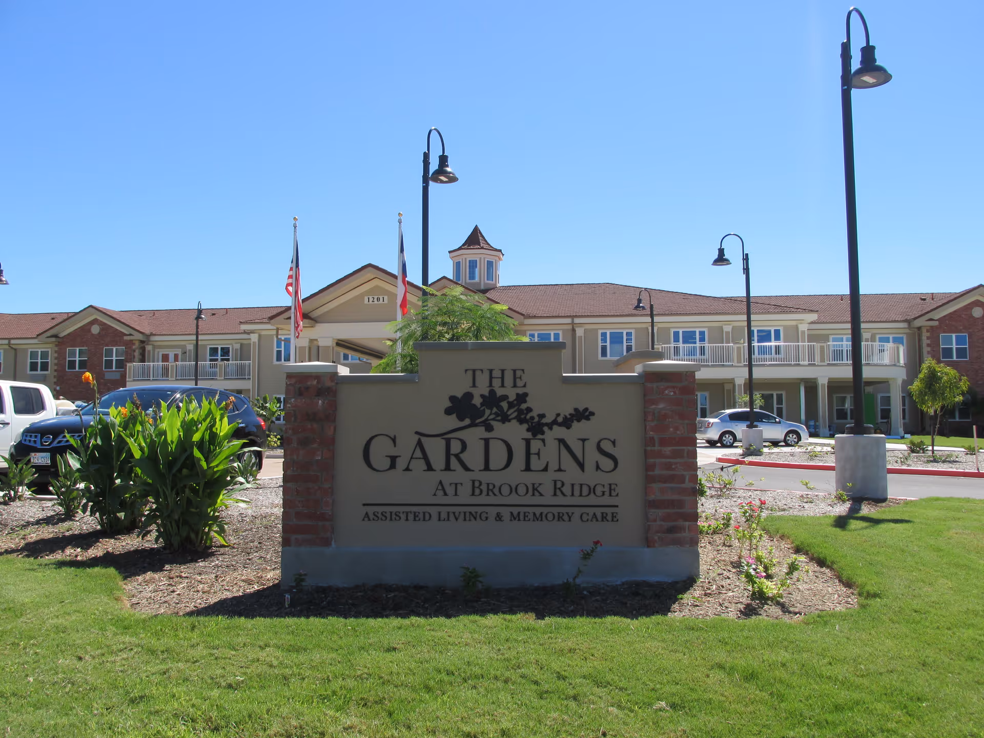 Exterior view of The Gardens at Brook Ridge Assisted Living & Memory Care facility with a large sign in the foreground displaying the facility's name. The building is two stories with balconies, surrounded by landscaped greenery, and several cars are parked nearby under a clear blue sky.