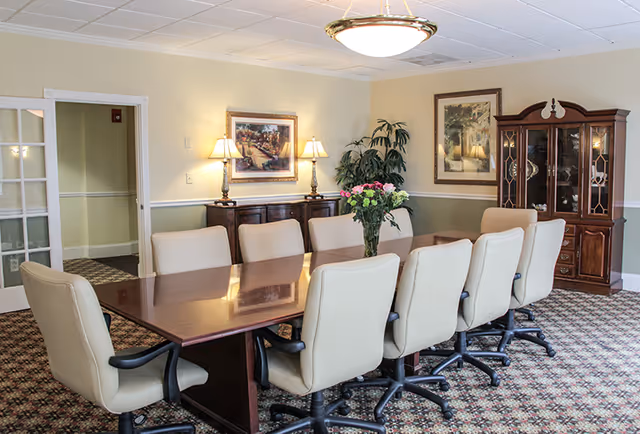 A formal dining/meeting room with a long wooden table surrounded by tan swivel chairs, a floral centerpiece, lamps, framed artwork, and a china cabinet.