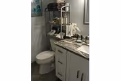 A small bathroom featuring a white toilet and a white vanity with a marble countertop. On the countertop are various toiletries and a small decorative item. Above the toilet is a metal shelving unit holding additional items. The walls are light-colored, and a framed picture is partially visible on the left wall.