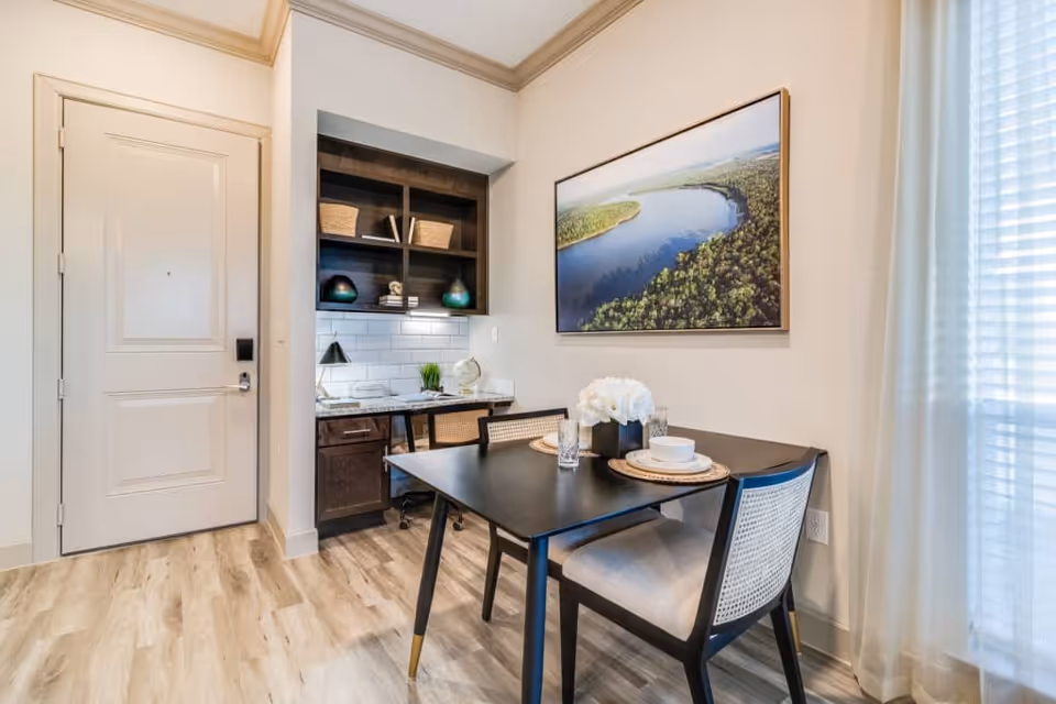 Small dining area with a black table set for two, woven-back chairs, a built-in desk and shelving next to an entry door, and a large landscape print on the wall.