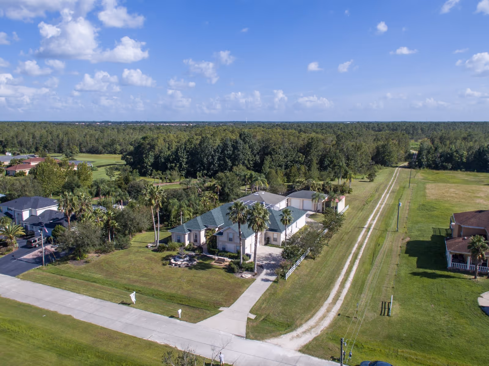 Aerial view of a single-story residential building with a green roof surrounded by palm trees and greenery. The building is situated on a large grassy lot with a paved driveway and a gravel road running alongside it. Other houses and dense forested areas are visible in the background under a partly cloudy blue sky.