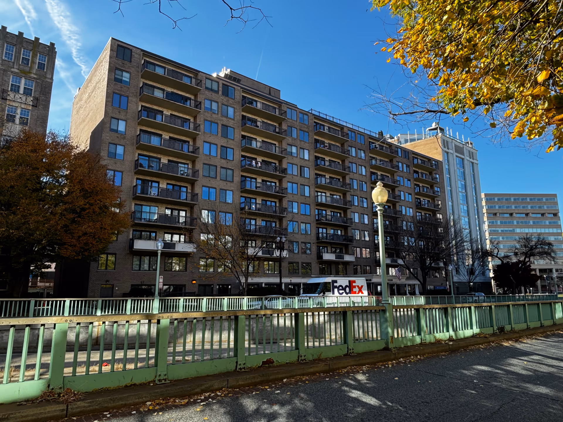 Exterior view of a multi-story brick building with balconies, identified as Thomas House Retirement Community, under a clear blue sky. There are trees with autumn leaves and a green metal fence in the foreground. A FedEx truck is parked in front of the building.