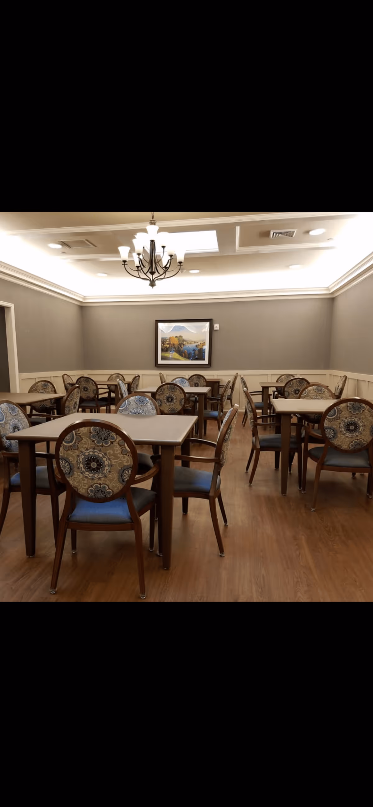 A dining room with multiple square tables and chairs arranged neatly. The chairs have patterned upholstery with blue and beige tones. The room has wooden flooring, gray walls with white wainscoting, a chandelier hanging from the ceiling, and a framed landscape painting on the far wall.