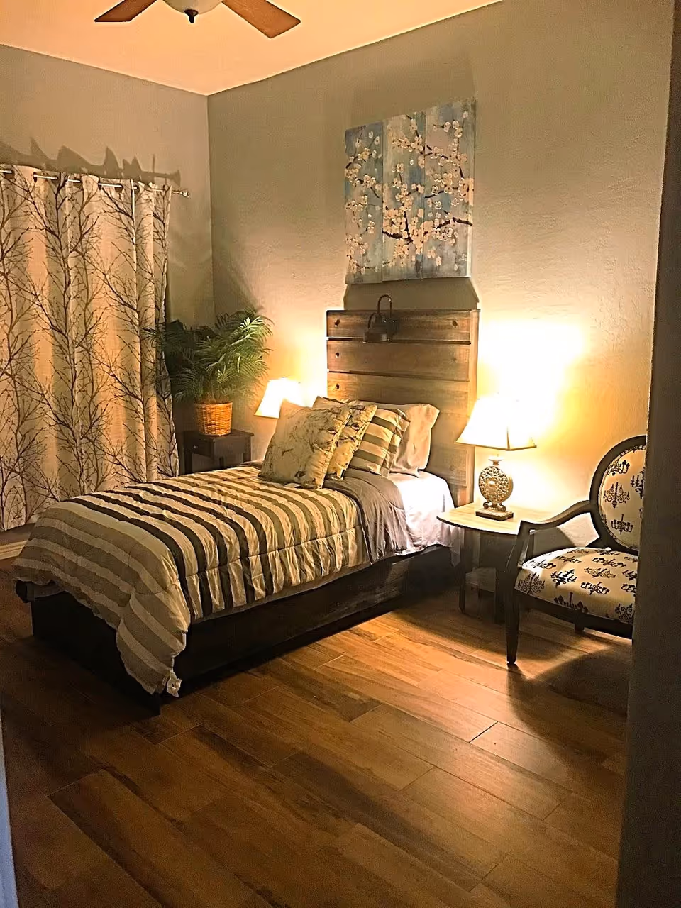 A cozy bedroom with a single bed featuring striped bedding and multiple pillows. There is a wooden headboard with a decorative lock, two bedside tables each with a lamp, a patterned armchair, a potted plant, and curtains with a tree branch design. The room has wooden flooring and a ceiling fan.