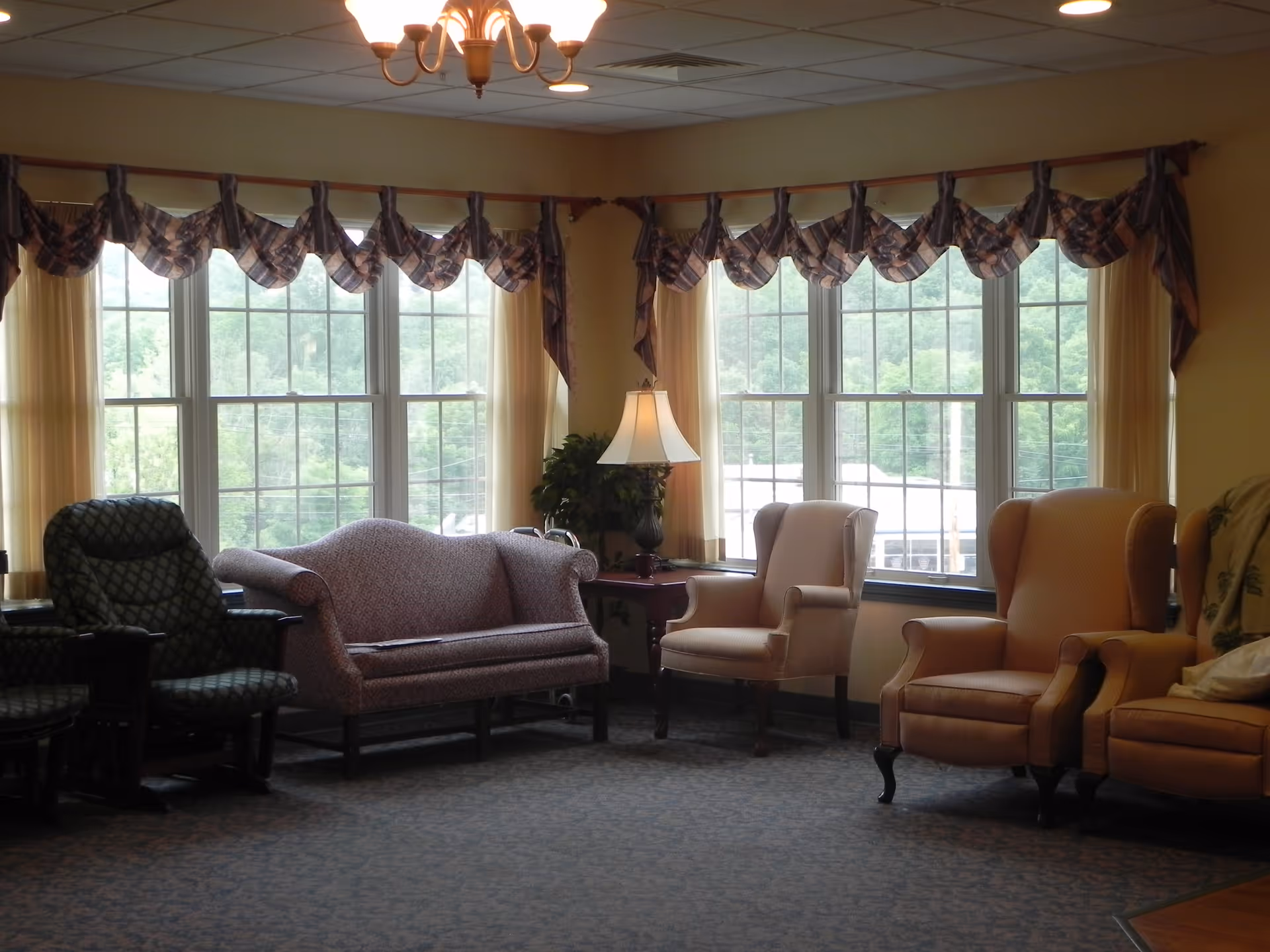 Sunlit common sitting area with upholstered armchairs and a loveseat arranged before large multi-pane windows.