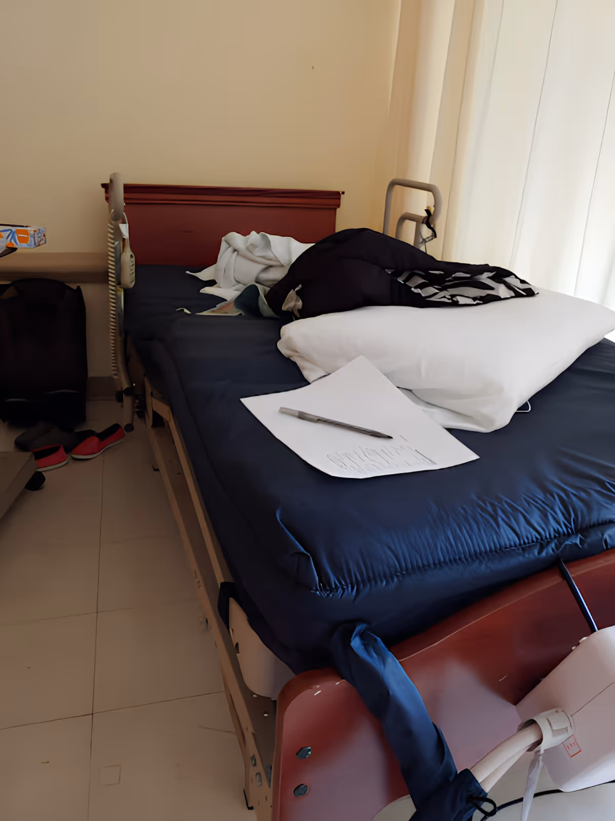 A hospital-style bed with a dark blue mattress and a white pillow, partially covered with a black and white blanket. There is a piece of paper and a pen on the bed. The bed has side rails and is positioned next to a window with white curtains. A pair of red slippers and a black bag are on the floor beside the bed.