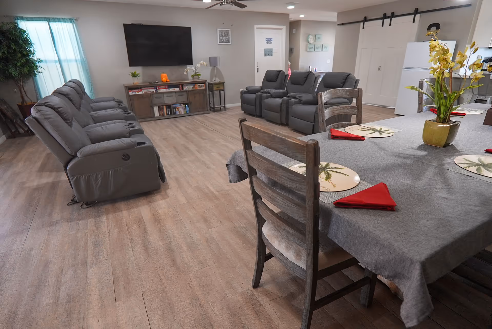 Spacious communal living area with a dining table and place settings in the foreground and rows of gray recliners facing a wall-mounted TV and media console in the background.
