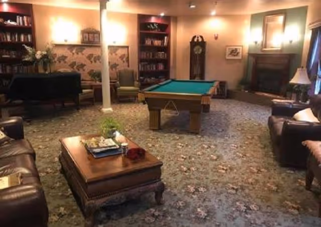 A cozy living room with a pool table in the center, surrounded by leather sofas and armchairs. There is a wooden coffee table with books and a plant on it. The room features a grandfather clock, a fireplace, bookshelves, and warm lighting from wall sconces and lamps. The carpet has a floral pattern.