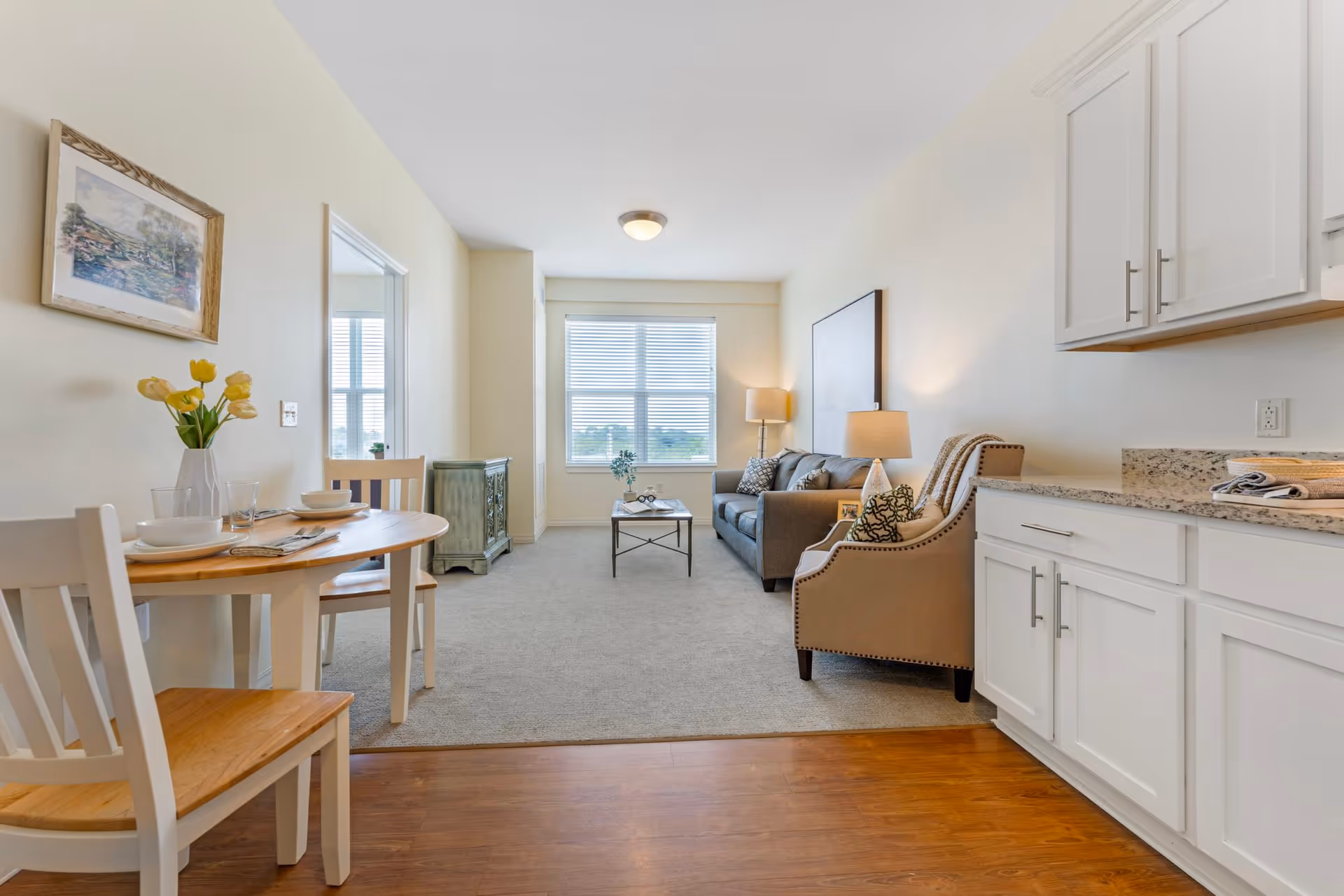 A bright and clean living area in a senior living facility featuring a small dining table with two chairs and place settings, a cozy sitting area with a sofa, armchair, coffee table, and two lamps, and a kitchen counter with white cabinets and granite countertops. A large window at the far end lets in natural light, and a framed picture hangs on the wall.