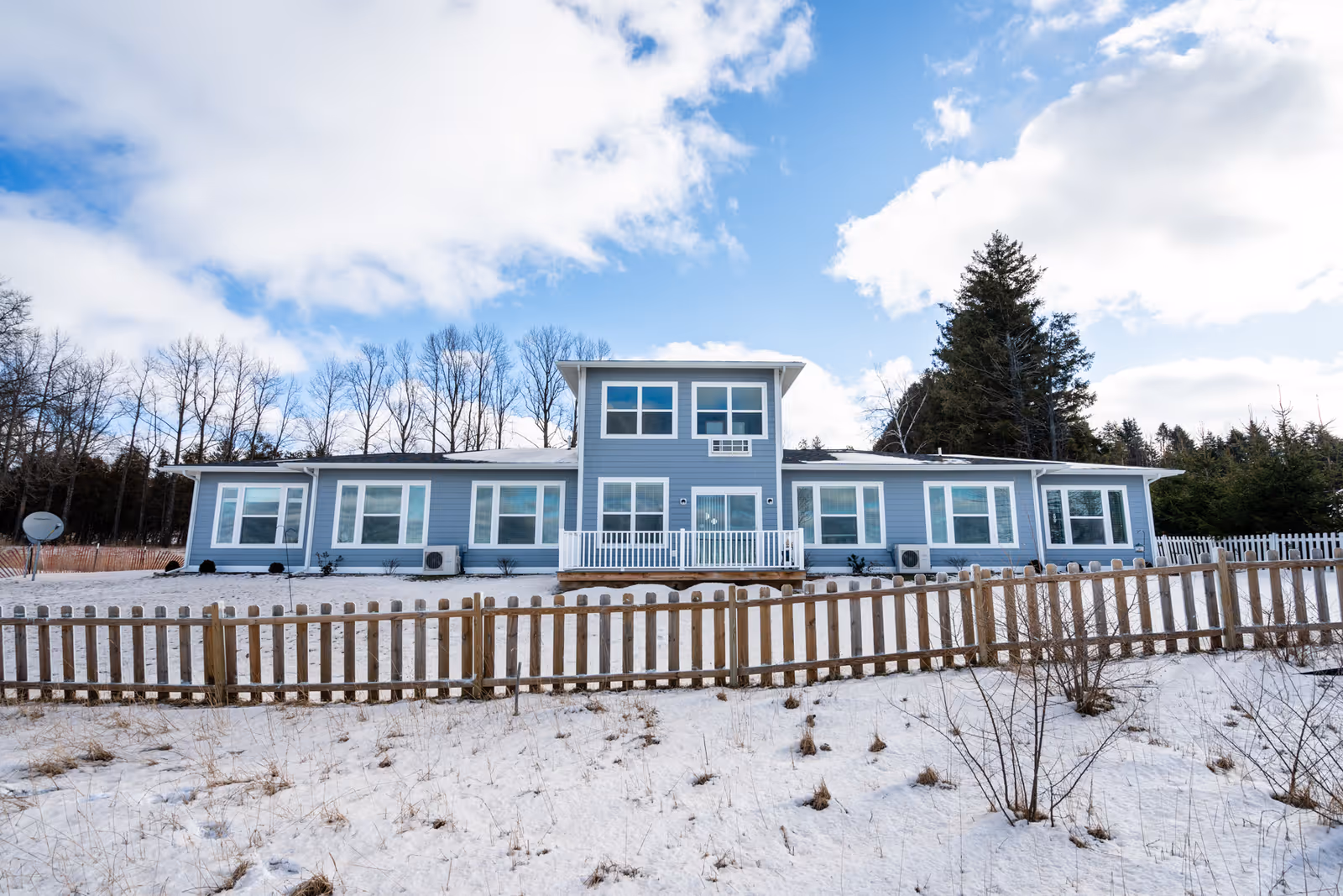 Exterior view of a single-story blue senior living facility building with multiple large windows, a small porch with white railing, surrounded by a wooden fence and snow-covered ground under a partly cloudy sky.