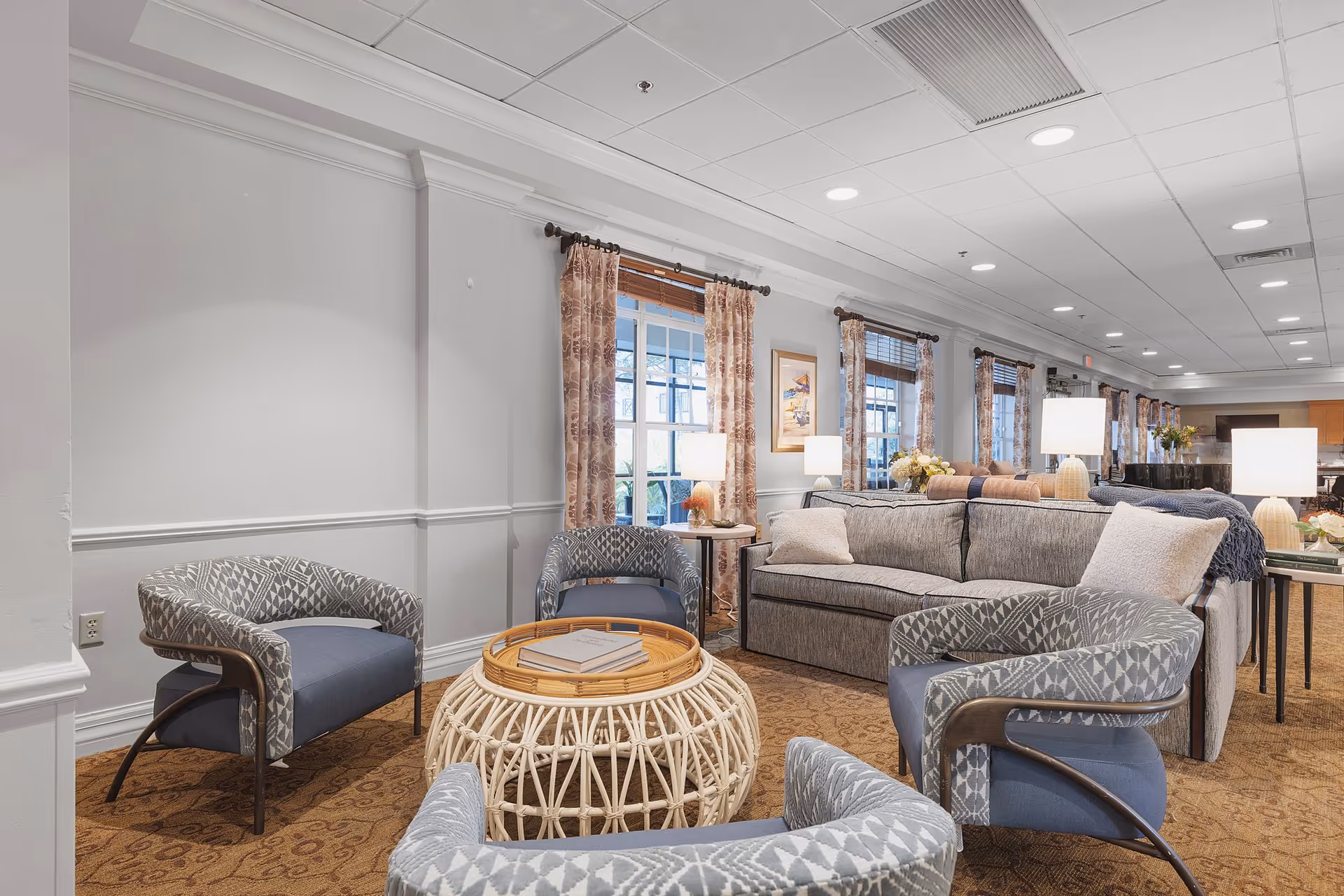 A cozy living room area in a senior living facility featuring a gray sofa with pillows, four patterned armchairs arranged around a round wicker coffee table with books on top, multiple table lamps, and windows with floral curtains letting in natural light.