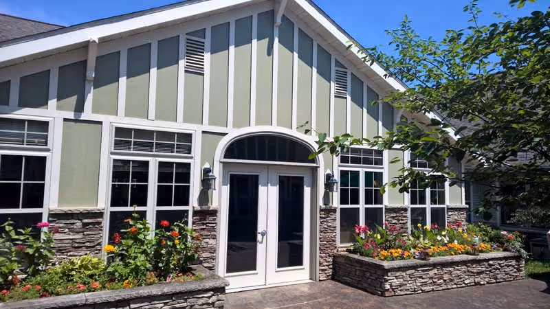 Exterior view of a building with light green and white vertical paneling and stone accents. The building has a double door entrance with an arched window above it, flanked by two wall-mounted lanterns. There are flower beds with colorful flowers on either side of the entrance, and a tree partially visible on the right side under a clear blue sky.