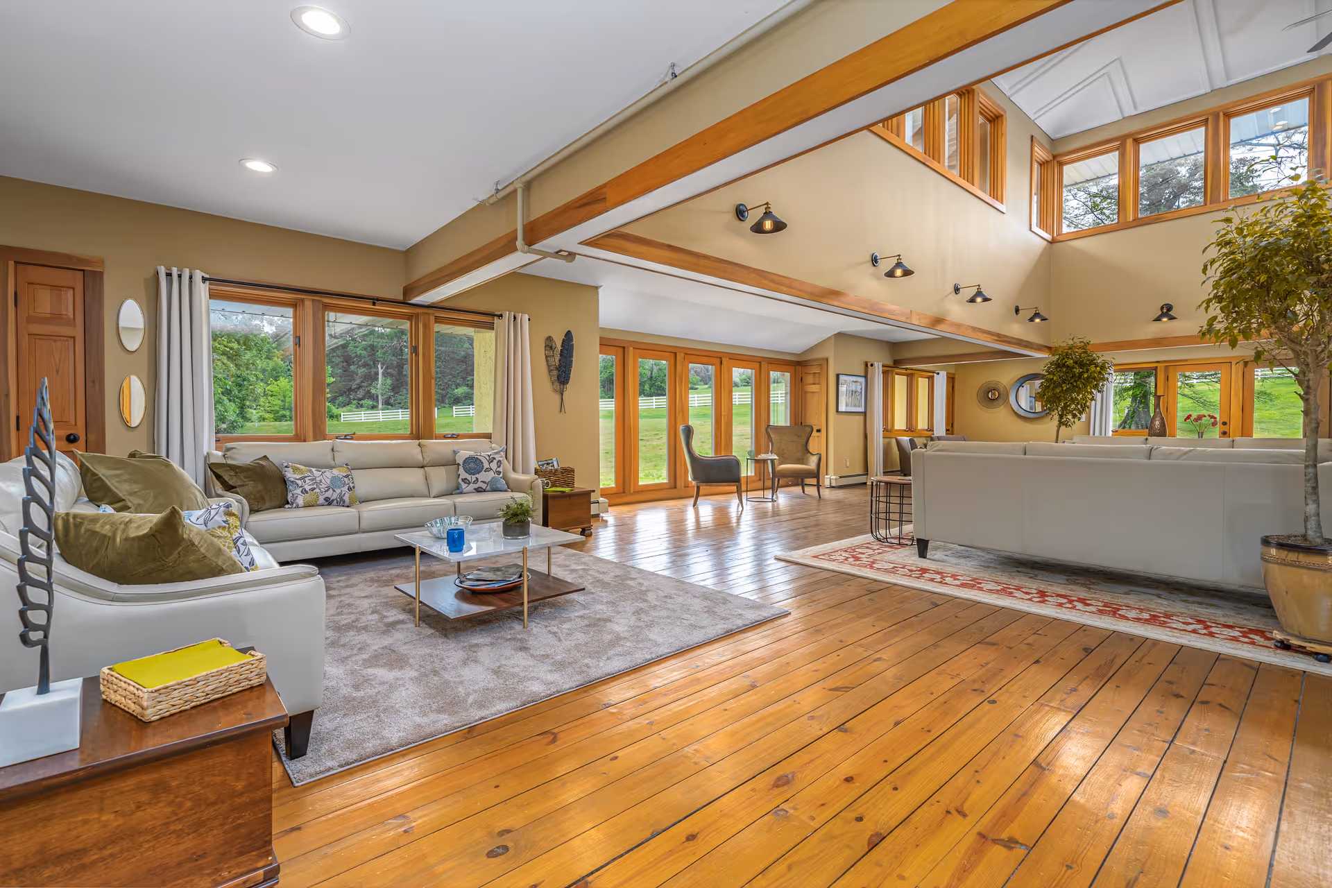 Spacious living room with wooden floors, beige walls, and large windows offering a view of green outdoor scenery. The room features two white sofas with decorative pillows, a coffee table on a carpet, two armchairs near glass doors, and indoor plants. The ceiling is high with exposed wooden beams and multiple wall-mounted lights.