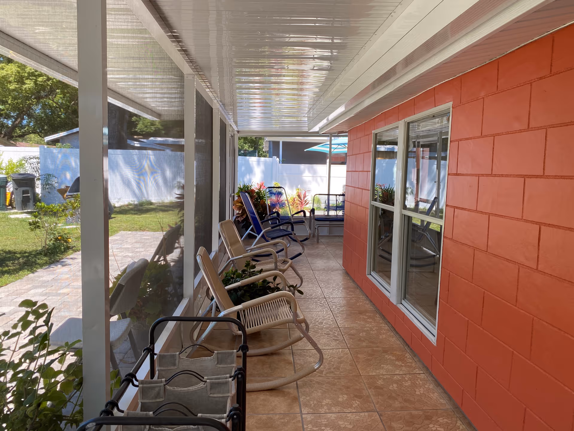 A covered outdoor patio area with several chairs lined up along a red brick wall. The patio has tiled flooring and is enclosed with mesh screens. There are plants and a small garden visible outside the screened area, along with a white fence and some outdoor furniture including an umbrella.