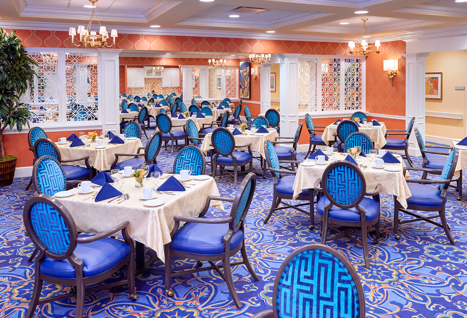 A bright and elegant dining room with multiple tables covered in cream-colored tablecloths, each set with white cups, plates, silverware, and blue folded napkins. The chairs have dark wooden frames with blue patterned upholstery. The room features a blue and purple patterned carpet, coral-colored walls with white trim, decorative white lattice panels, and several chandeliers providing warm lighting.