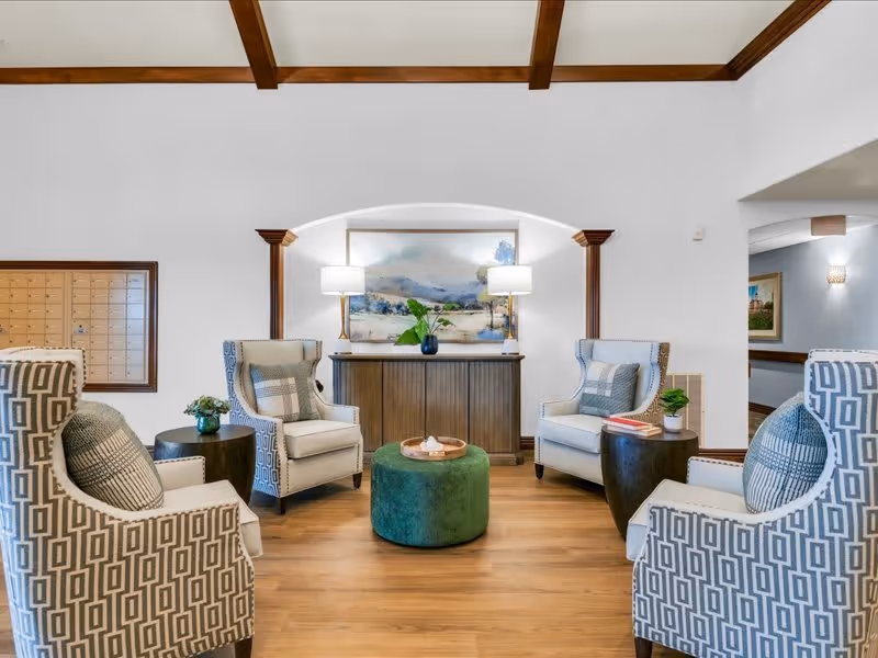 A cozy seating area in a senior living facility with four patterned armchairs arranged around a green round ottoman. Behind the chairs is a wooden console table with two lamps and a large landscape painting. The room has wooden beams on the ceiling and light wood flooring.