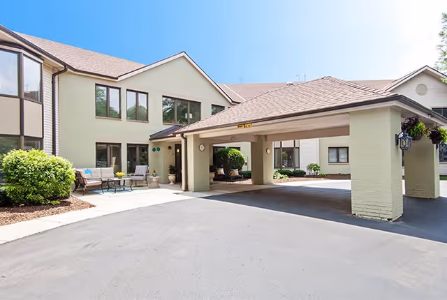 Exterior view of a senior living facility building with a covered entrance driveway. The building has beige walls, multiple windows, and a seating area with chairs and a table near the entrance. There are some green bushes and plants around the building under a clear blue sky.