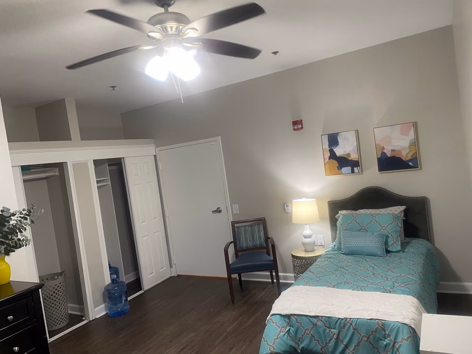 Well-lit single-occupancy bedroom with a teal patterned bed, nightstand and lamp, chair, ceiling fan, and open closet.