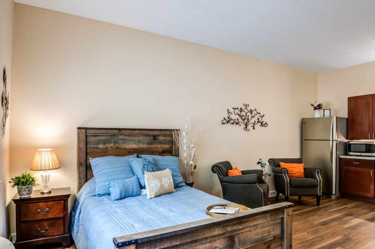 A cozy studio apartment with a wooden bed frame and blue bedding, a wooden nightstand with a lamp and plant, two dark upholstered armchairs with orange pillows, a small side table with flowers, and a kitchenette area with a stainless steel refrigerator, microwave, and wooden cabinets.