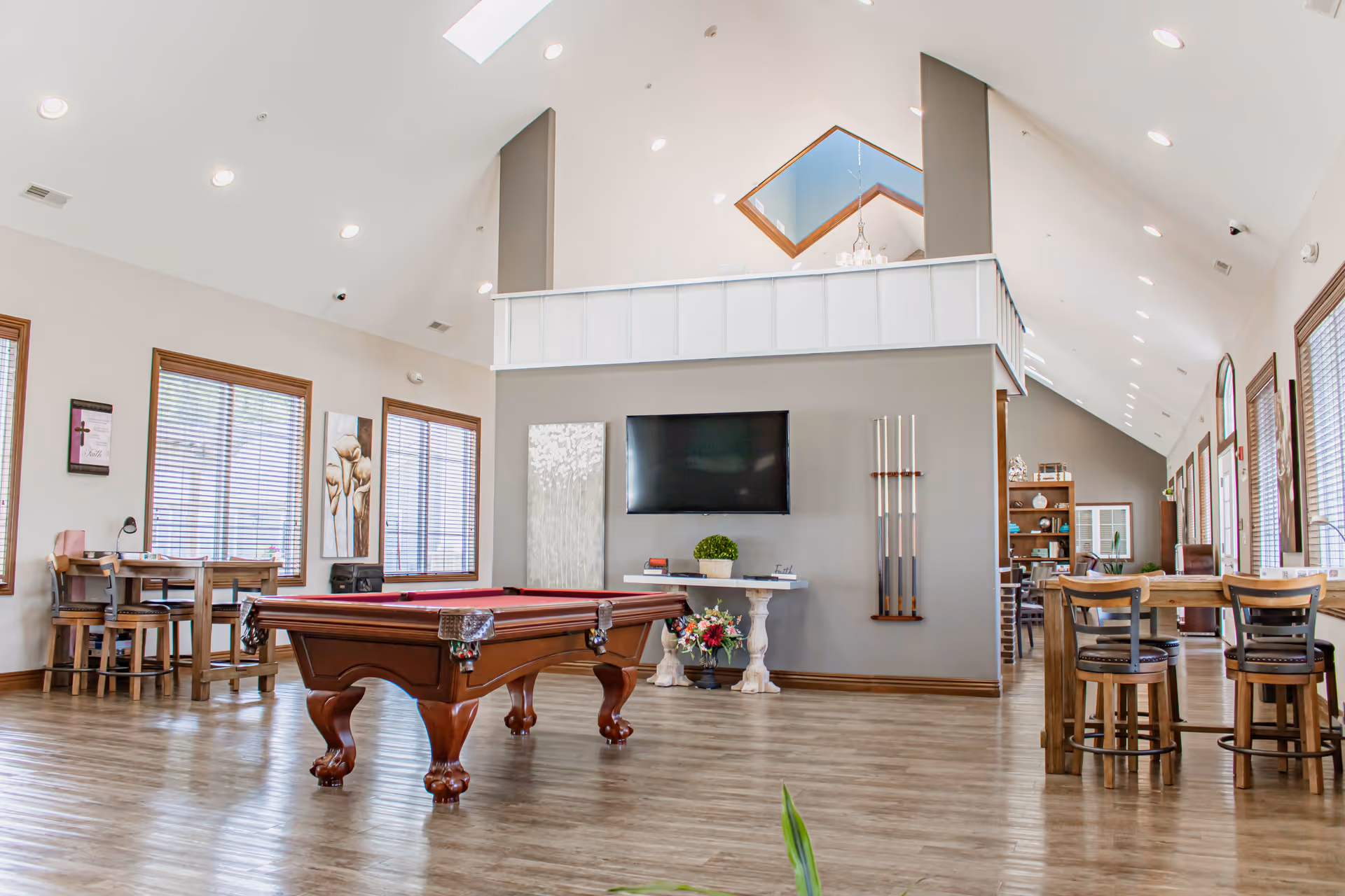 A spacious and well-lit common area with a pool table in the center, a wall-mounted TV, and pool cues on the wall. The room features high vaulted ceilings with skylights, large windows with wooden blinds, and wooden flooring. There are multiple seating areas with tables and chairs along the sides of the room, and decorative plants and artwork on the walls.