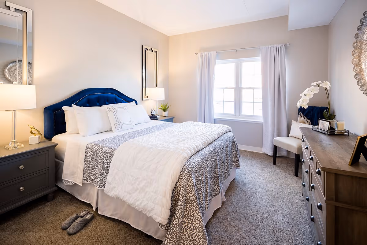 A bright and cozy bedroom with a large bed featuring a blue upholstered headboard, white and patterned bedding, and multiple pillows. There are two nightstands with lamps on either side of the bed, a pair of slippers on the carpeted floor, a window with white curtains letting in natural light, a wooden dresser with decorative items including a white orchid, and a chair in the corner.
