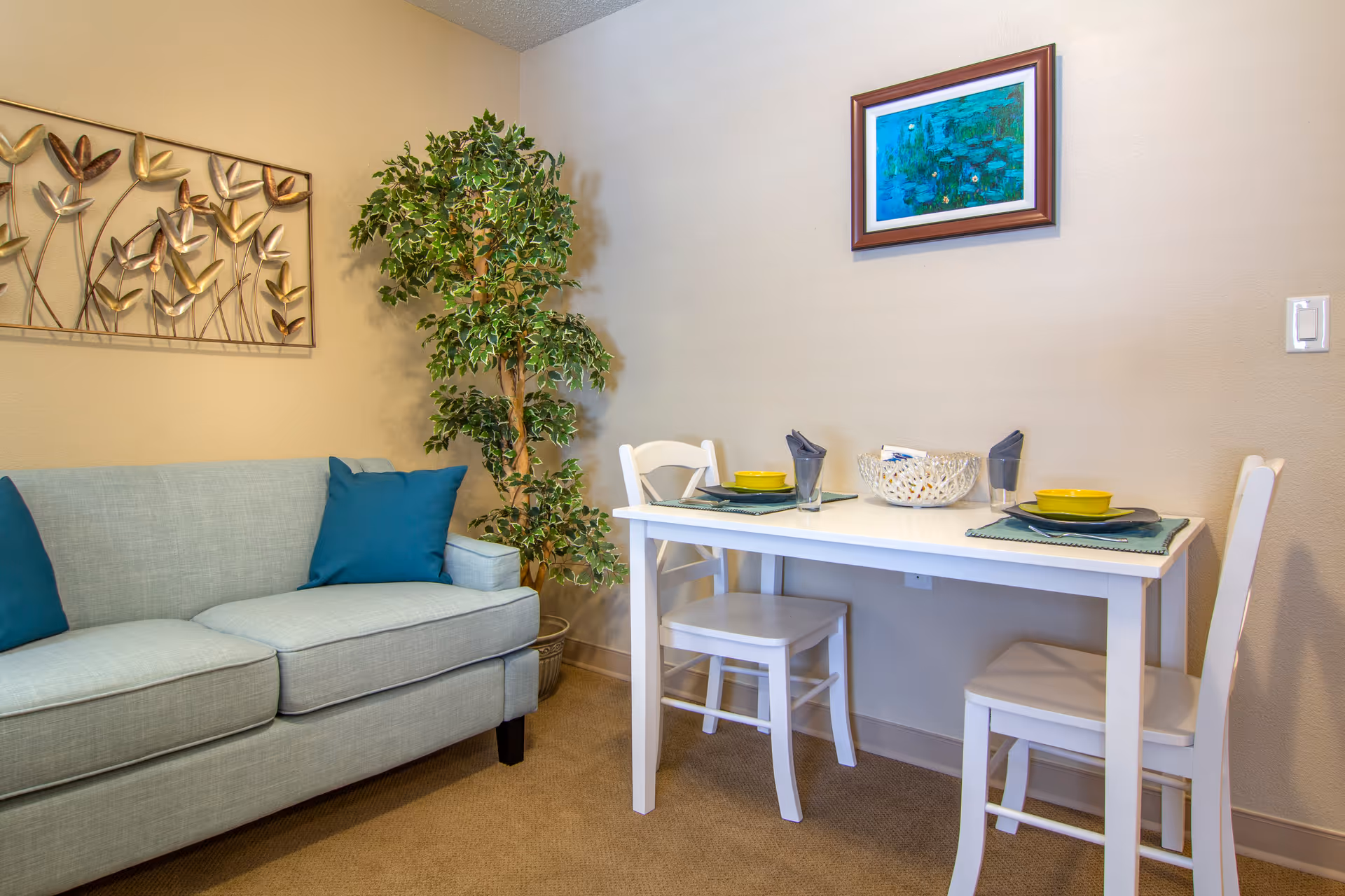 A small dining area with a white table set for two with yellow bowls, gray napkins, and glasses. Next to the table is a light blue sofa with two blue cushions. A tall green potted plant is placed in the corner, and a framed painting with blue and green tones hangs on the beige wall above the table.
