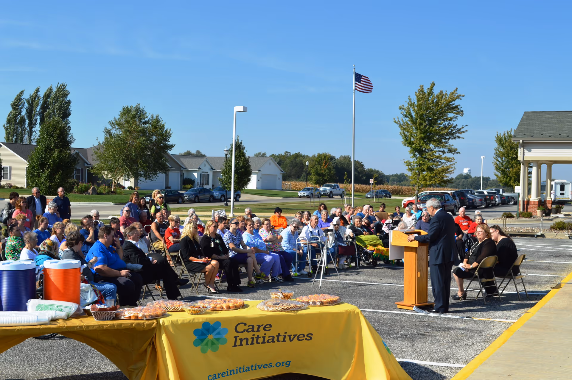 Outdoor ceremony with residents and guests seated listening to a speaker at a podium beside a table draped with a yellow 'Care Initiatives' tablecloth and an American flag in the background.