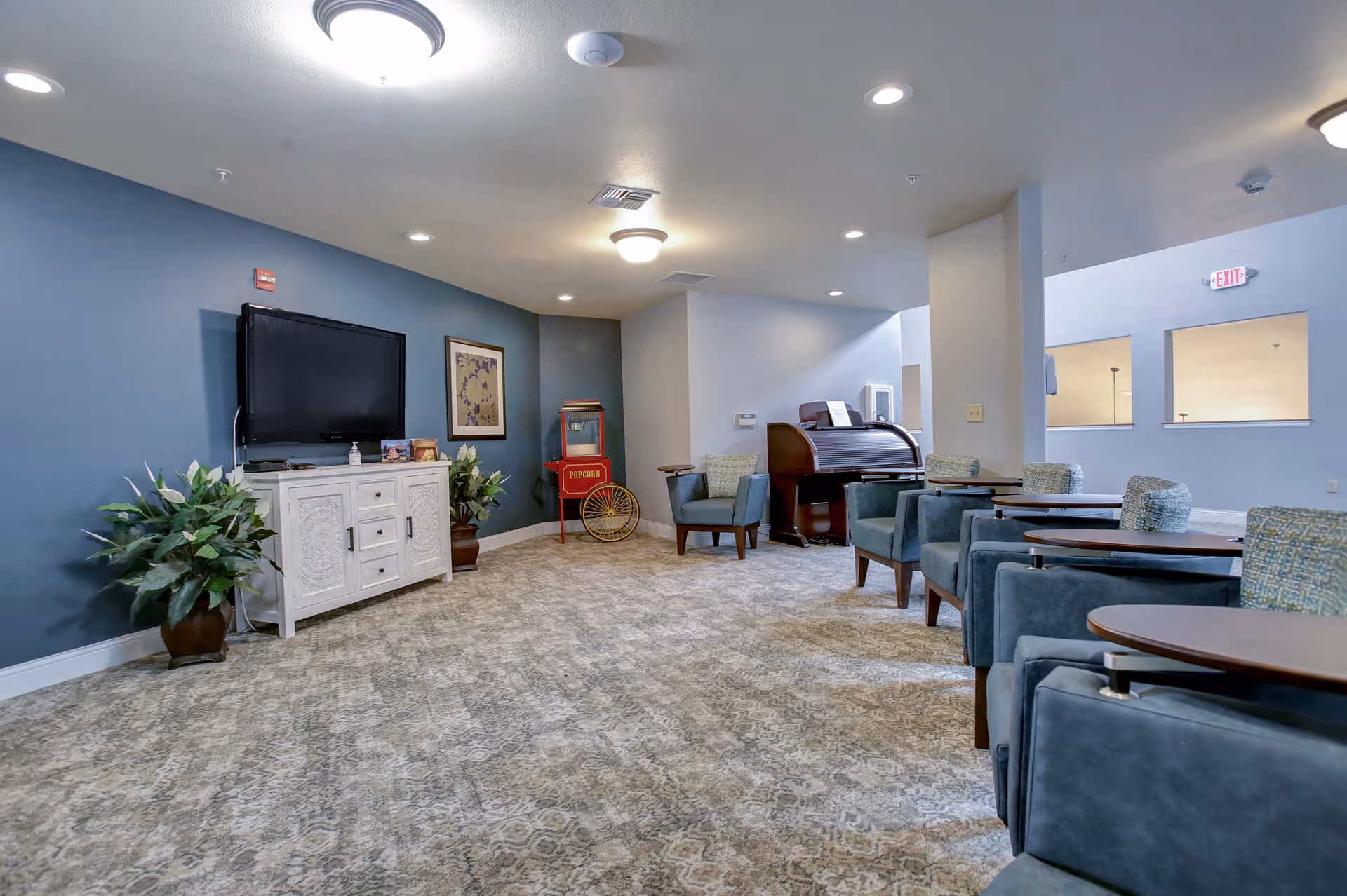 A senior living facility common area with blue and white walls, patterned carpet, and several blue upholstered chairs with attached wooden tables. There is a white cabinet with a flat-screen TV mounted above it, two potted plants on either side, a red popcorn machine in the corner, and a wooden roll-top desk with a chair. The room is well-lit with ceiling lights.