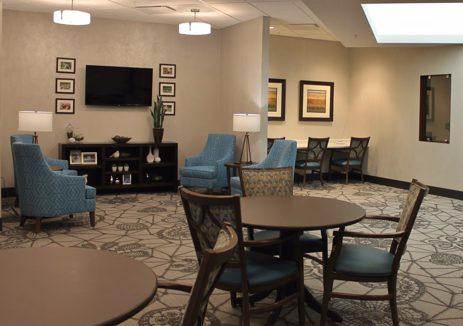 Communal lounge with round tables, upholstered chairs, a wall-mounted TV, and framed artwork.