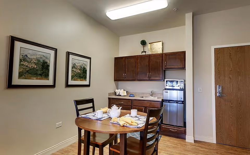 A small dining area in a senior living facility with a round wooden table set for tea, including a teapot, cups, and napkins. The room features light-colored walls, two framed landscape paintings, wooden cabinets with a small sink, a microwave, and a mini refrigerator. There is a wooden door on the right side of the image.