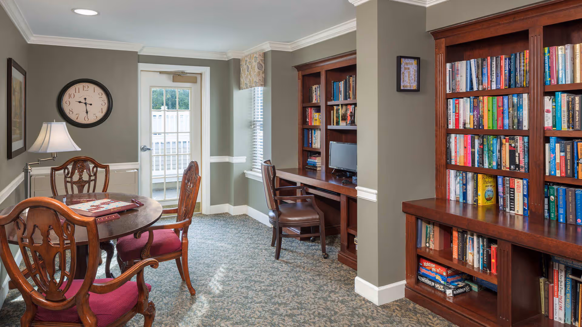 A cozy room with a round wooden table and four chairs with red cushions, a table lamp, a wall clock, a door with a window, a window with blinds, and built-in wooden bookshelves filled with books and board games. There is also a wooden desk with a chair and a small computer monitor.