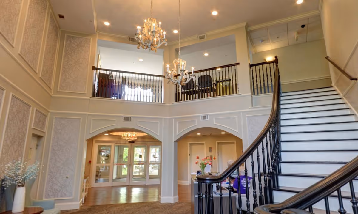 Interior view of a senior living facility lobby with a grand staircase on the right, decorative wall panels, two chandeliers hanging from the ceiling, and a balcony railing on the upper floor. There is a small table with flowers near the staircase and double glass doors leading outside in the background.