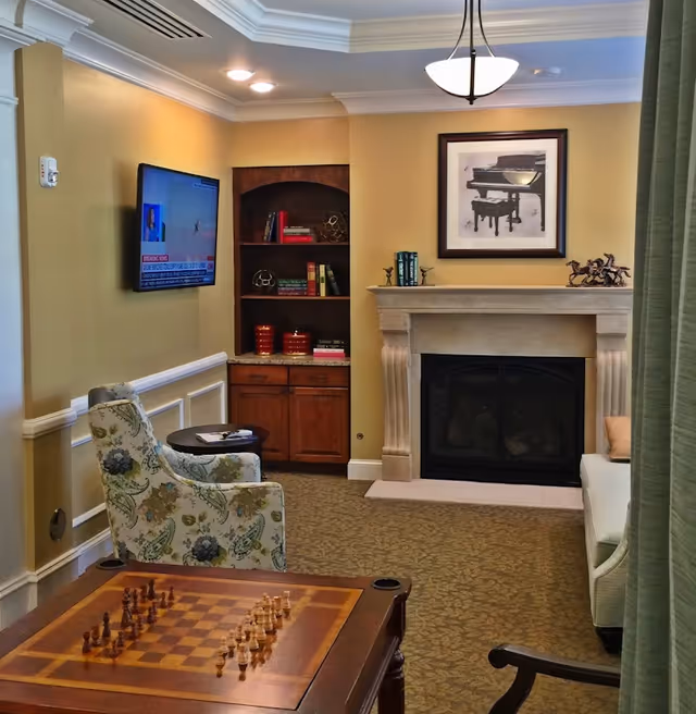 A cozy senior living room with a floral armchair, a chess table with a chessboard set up, a wall-mounted TV showing a news channel, a built-in wooden bookshelf with books and decorative items, a fireplace with a framed picture of a piano above it, and a beige sofa partially visible behind a green curtain.