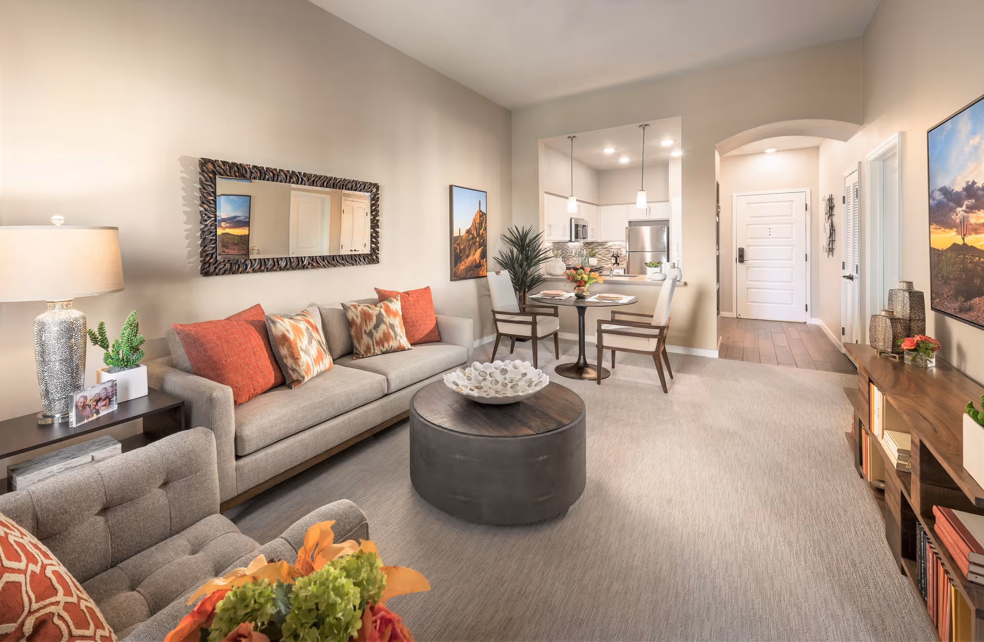 A bright and modern living room with a beige sofa adorned with red and patterned cushions, a round dark coffee table with a decorative bowl, a side table with a lamp and plant, and a small dining area with a round table and two chairs. The room has light-colored walls, carpeted floor, and an open view into a kitchen with stainless steel appliances. Artwork and a large mirror decorate the walls.