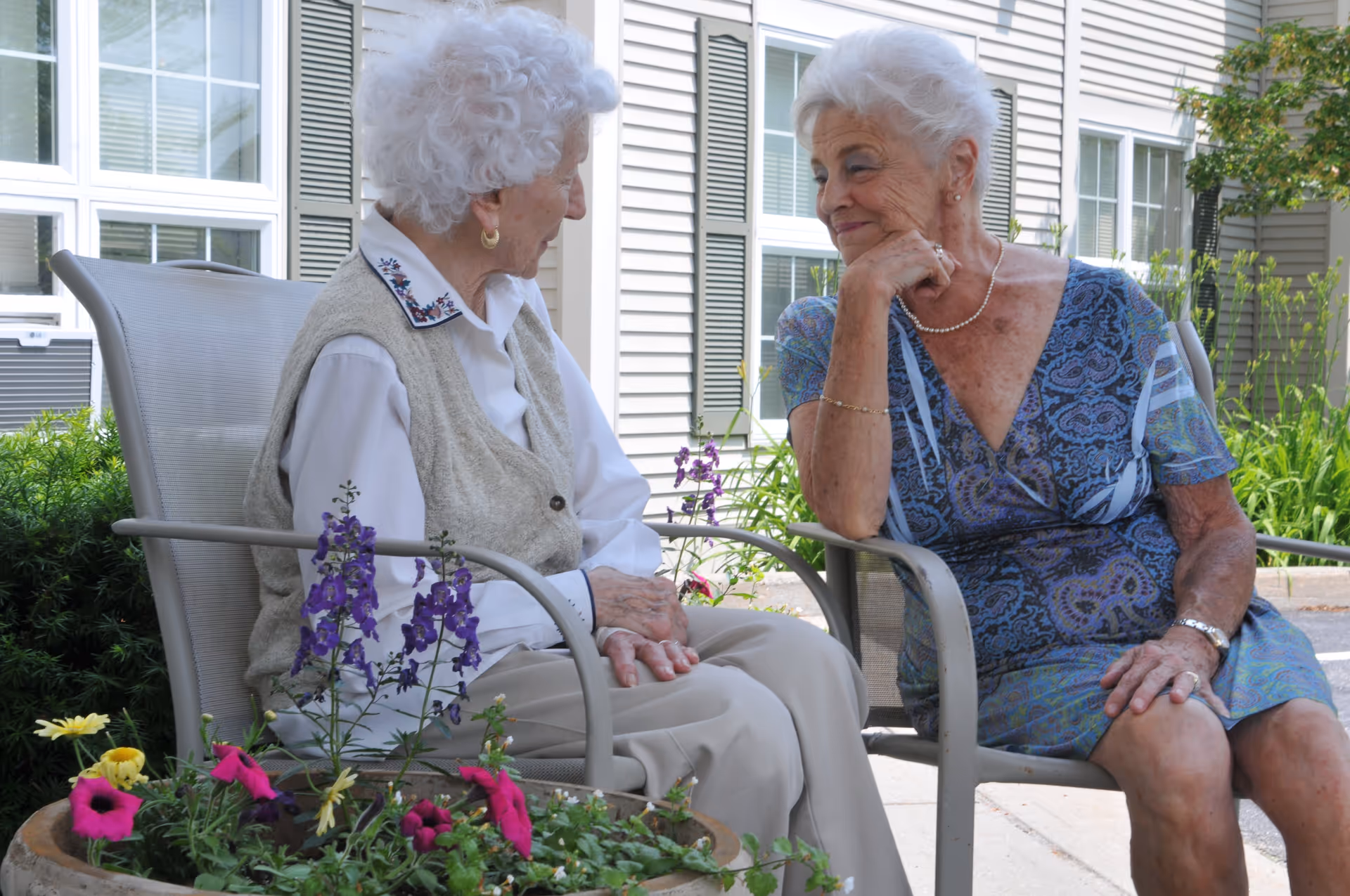 Two elderly women sitting outdoors on patio chairs, engaged in a friendly conversation. They are surrounded by flowering plants and greenery, with a building featuring windows and siding in the background.
