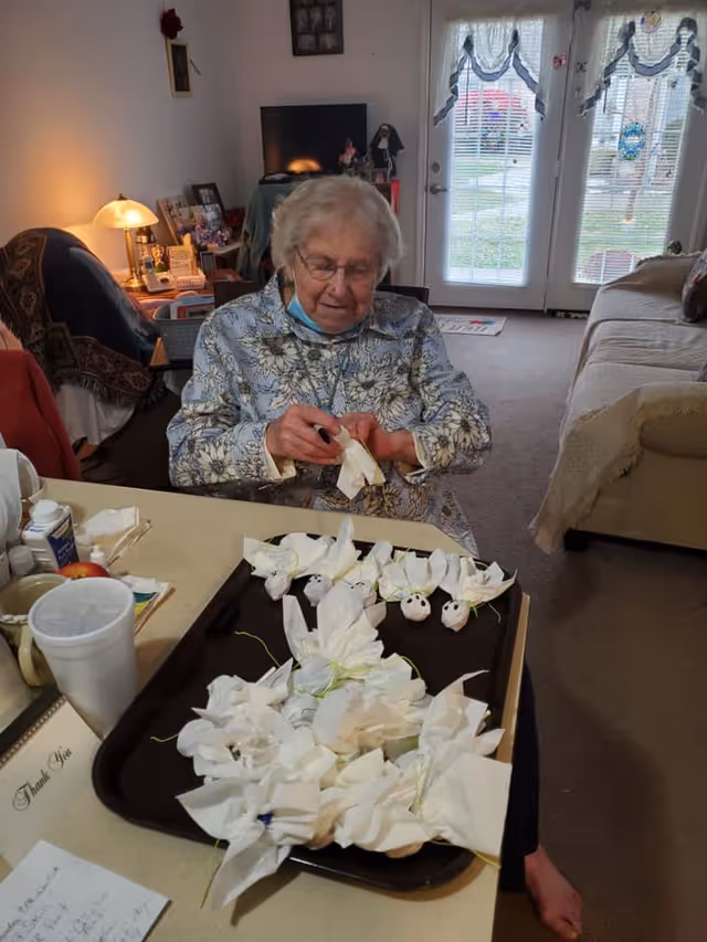 An elderly woman seated at a table in a living-room-style common area making paper crafts on a tray.