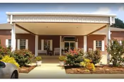 Front entrance of a senior living community building with a covered driveway, brick exterior, white columns, and landscaped bushes and flowers along the walkway.