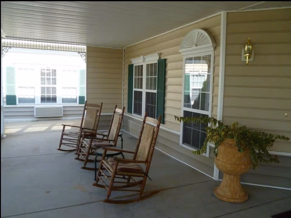 Covered outdoor patio area with three wooden rocking chairs arranged in a row facing outward. The patio has beige siding walls with white-trimmed windows and a large planter with green foliage on the right side. A brass wall-mounted light fixture is also visible on the wall.