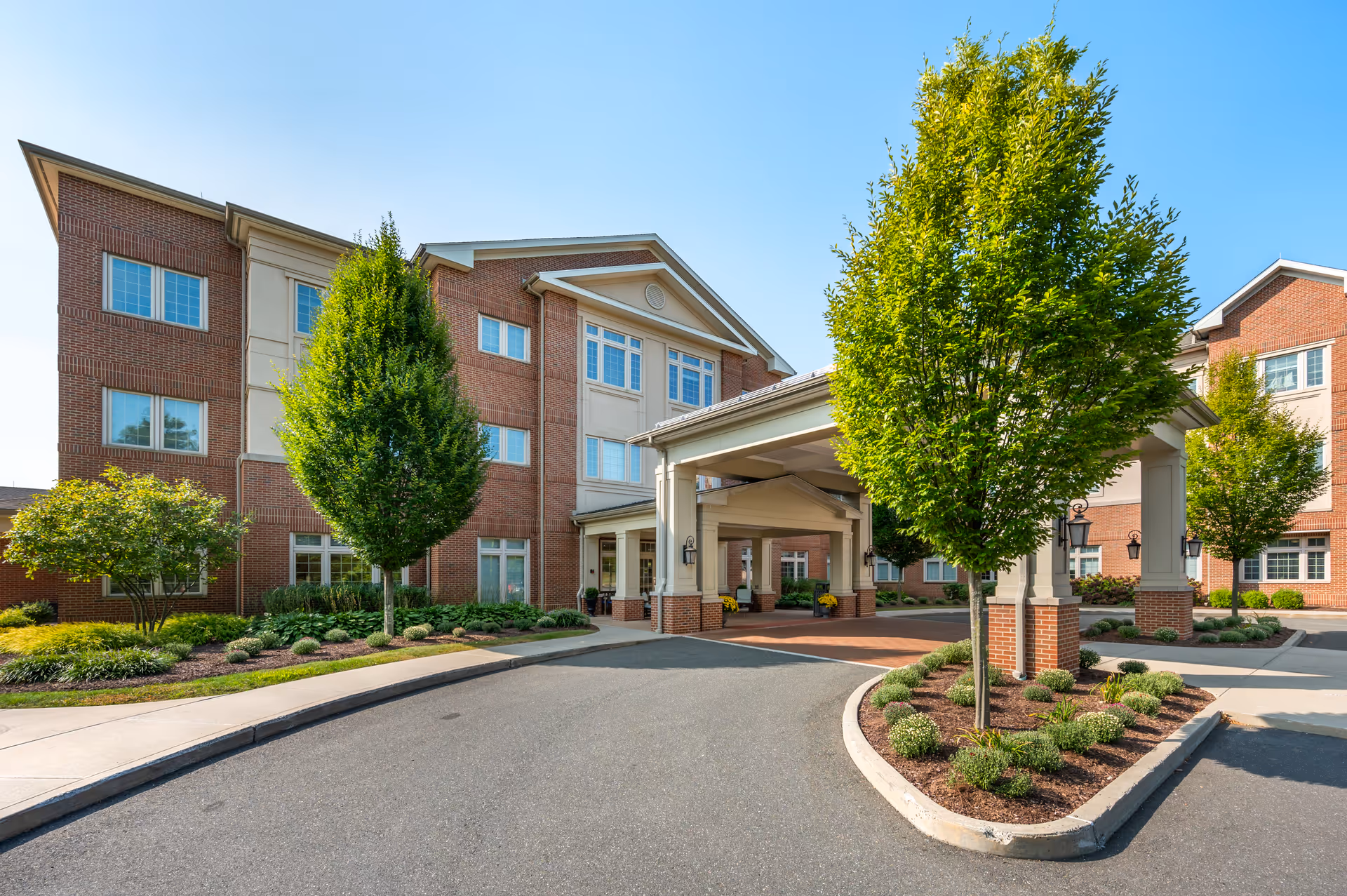 Front entrance of a multi-story brick senior living facility with a covered porte-cochère, landscaped driveway, and trees.
