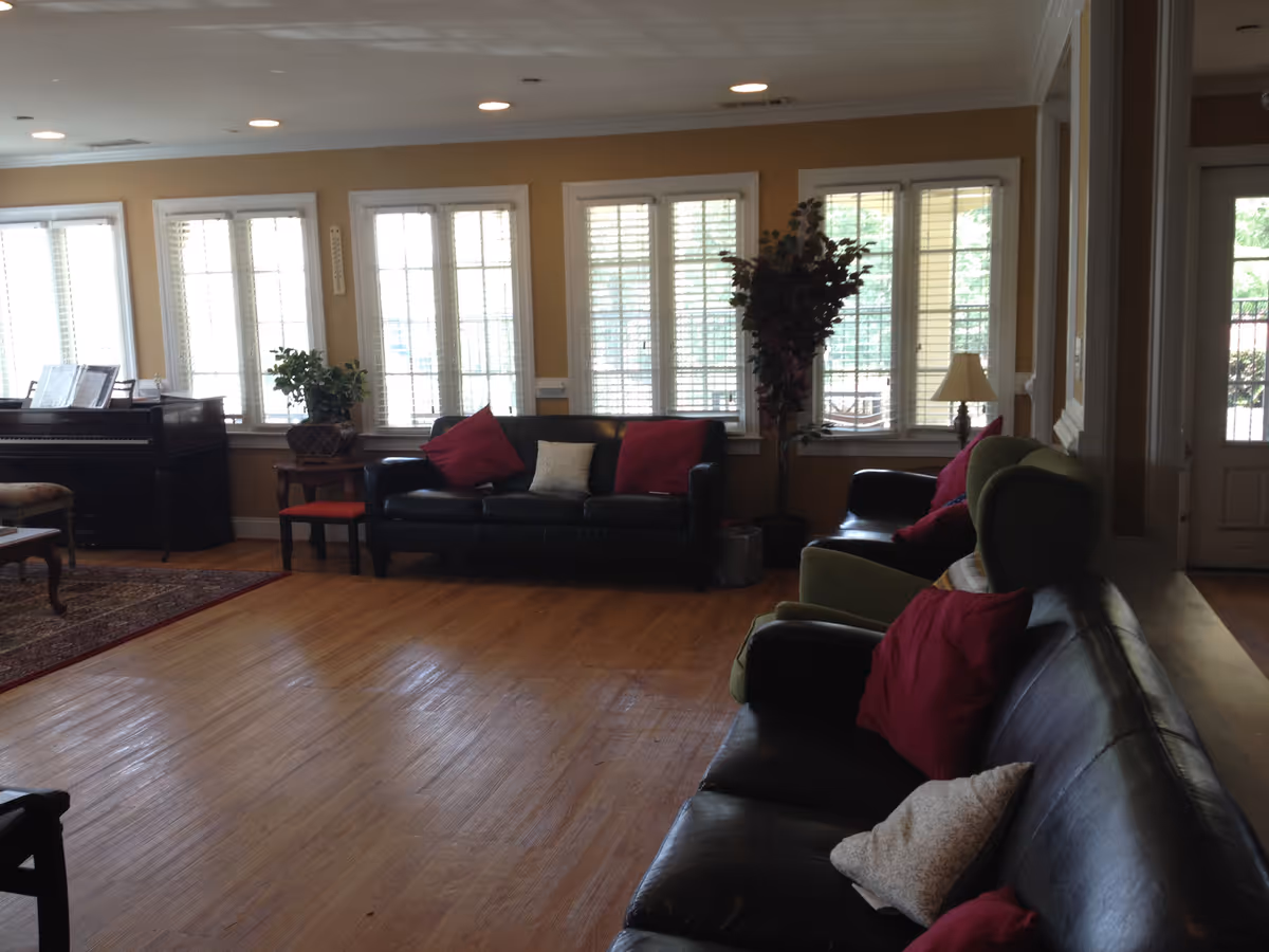 Sunlit common living room with sofas, armchairs, a piano, plants and large windows with blinds.