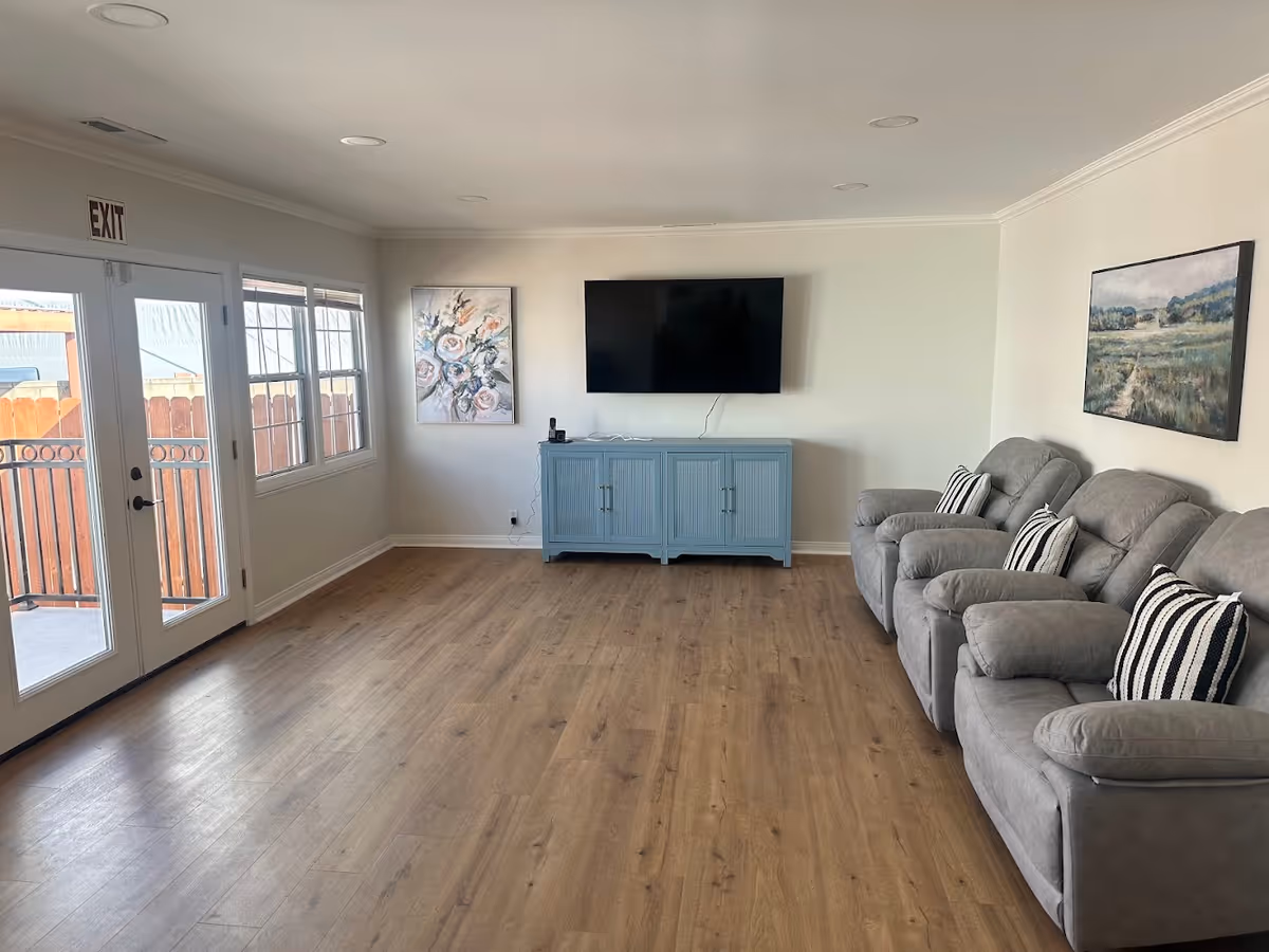 A living room with three gray recliner chairs with striped pillows on the right side, a blue cabinet with a flat-screen TV mounted above it on the far wall, and two paintings on the walls. There are double glass doors and a window on the left side letting in natural light, and the floor is wood.
