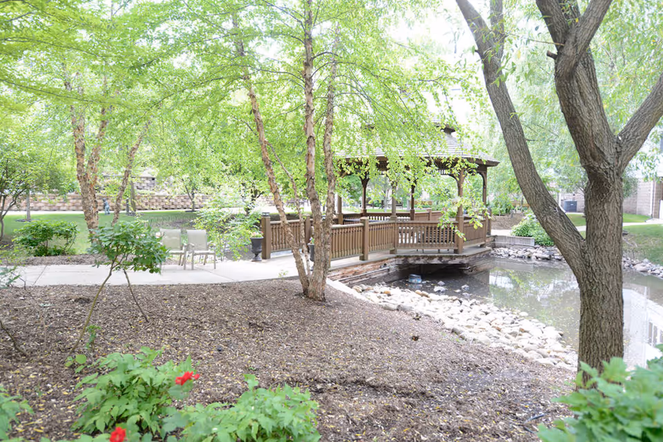 A peaceful outdoor garden area with green trees, shrubs, and a small pond. There is a wooden gazebo with a railing over the pond and a paved walkway with chairs nearby.